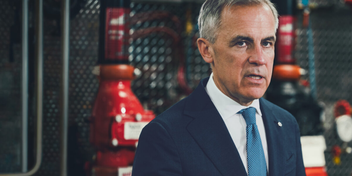 Prime Minister Mark Carney in a dark suit and blue tie, photographed in front of industrial pipefitting equipment at a Calgary union training facility.