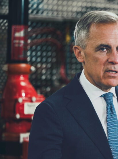 Prime Minister Mark Carney in a dark suit and blue tie, photographed in front of industrial pipefitting equipment at a Calgary union training facility.