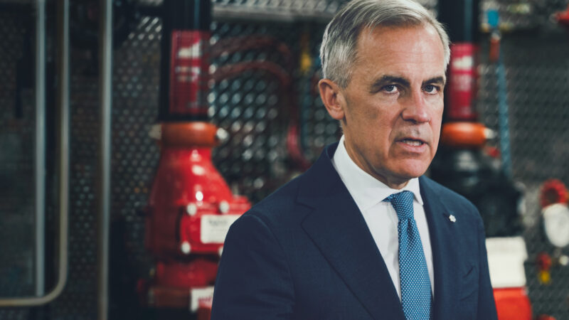 Prime Minister Mark Carney in a dark suit and blue tie, photographed in front of industrial pipefitting equipment at a Calgary union training facility.