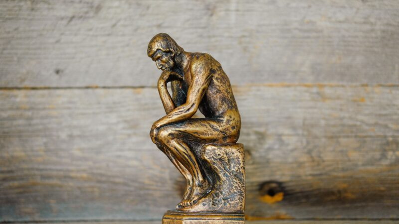 A bronze statue of a man in a pensive pose against a wooden background. Photo source: Quilia / Unsplash.
